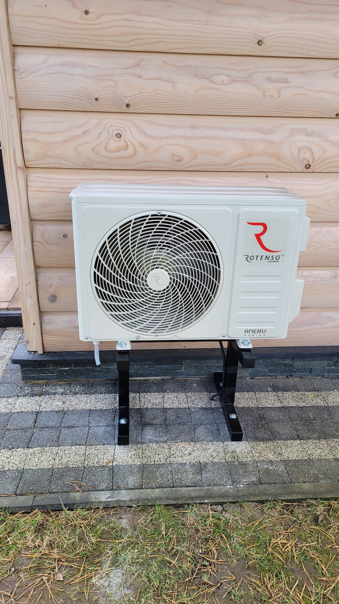 Biała jednostka zewnętrzna klimatyzatora Rotenso zamontowana na czarnym stelażu przy drewnianej ścianie budynku, widoczny fragment kostki brukowej i trawnika.
