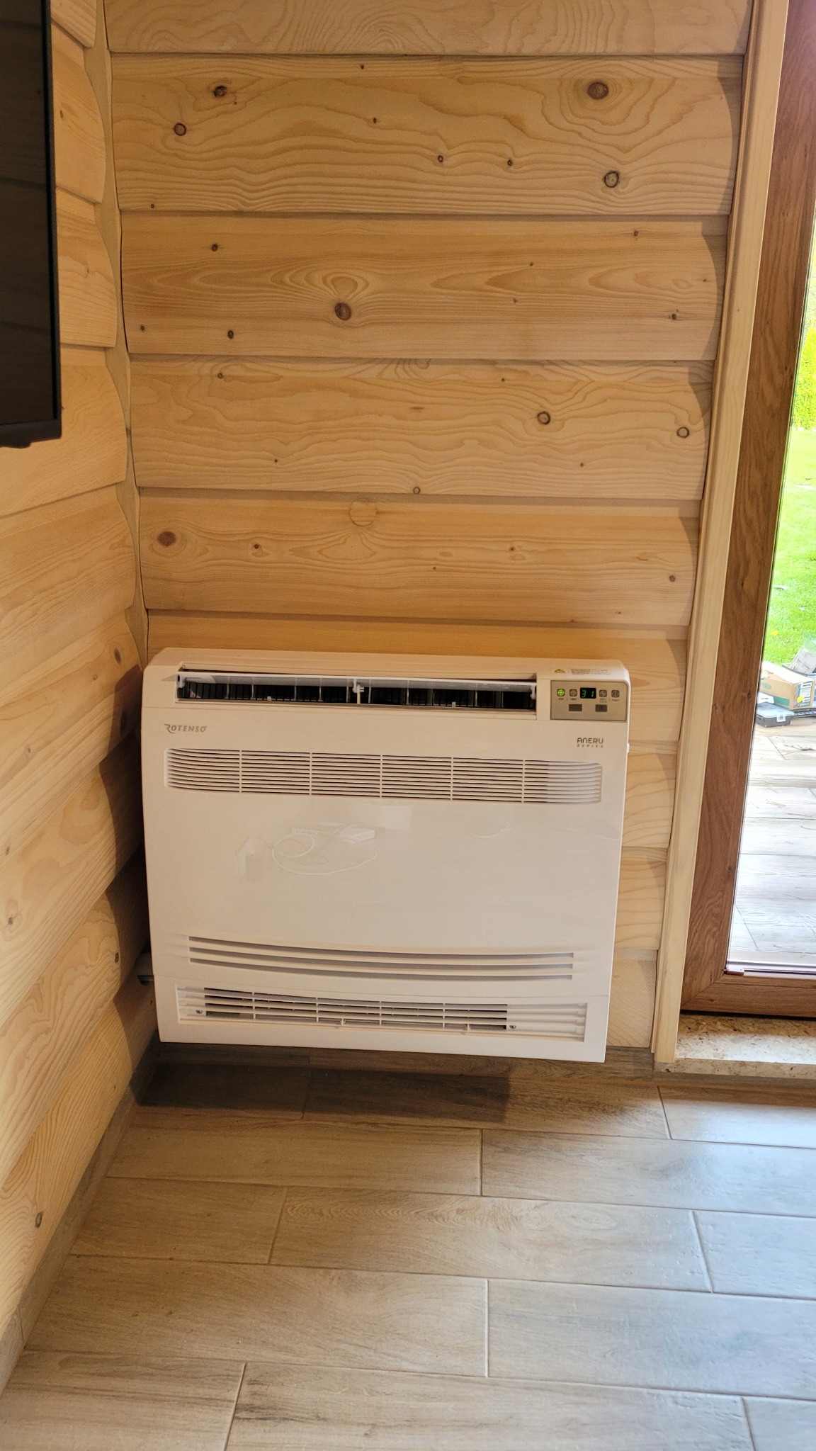 Biała jednostka wewnętrzna klimatyzatora podłogowego marki Rotenso, zamontowana na drewnianej ścianie w pomieszczeniu wykończonym w drewnie, z widoczną podłogą imitującą drewno i oknem w tle.