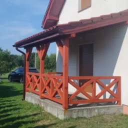 Zadaszony, drewniany ganek z balustradą w kształcie krzyży, pomalowany na czerwono-brązowo, przylegający do białego domu z brązowymi roletami i dachem, na tle zielonego trawnika i drzew.