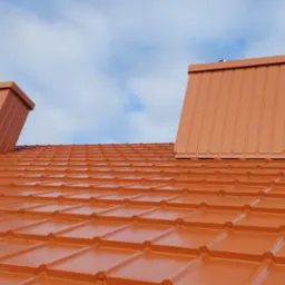 Pomarańczowa dachówka ceramiczna z dwoma kominami w kolorze, na tle błękitnego nieba z chmurami.