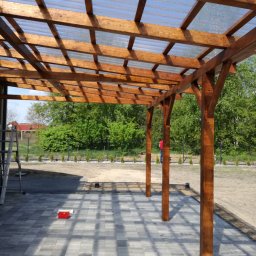 Usługi dekarsko-ciesielskie - Drewniana konstrukcja z przezroczystym zadaszeniem, widoczne słupy, fragment kostki brukowej, drabina i osoba w oddali na tle zieleni.