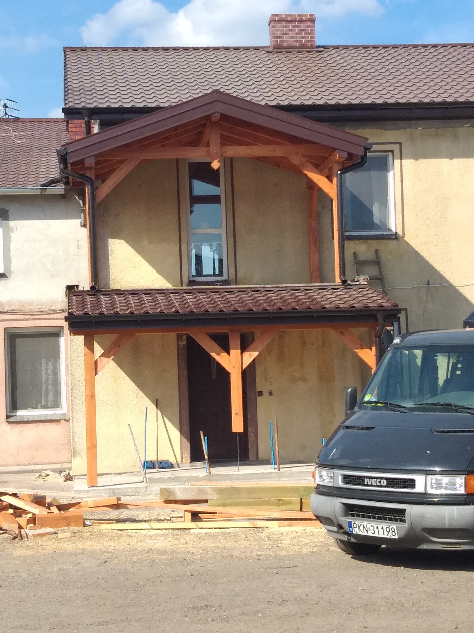 Drewniana konstrukcja z zadaszeniem nad wejściem do domu z brązową dachówką, widoczny fragment dachu budynku, zaparkowany samochód dostawczy i materiały budowlane na ziemi.