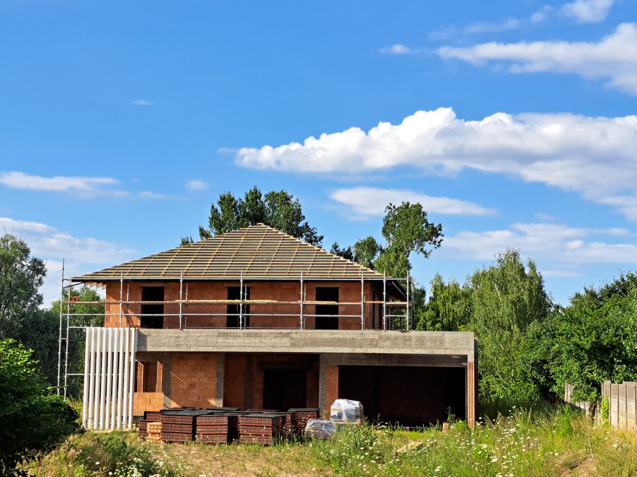 Dom w trakcie budowy z częściowo ukończoną konstrukcją dachu, widoczne rusztowania i materiały budowlane, na tle błękitnego nieba z białymi obłokami.