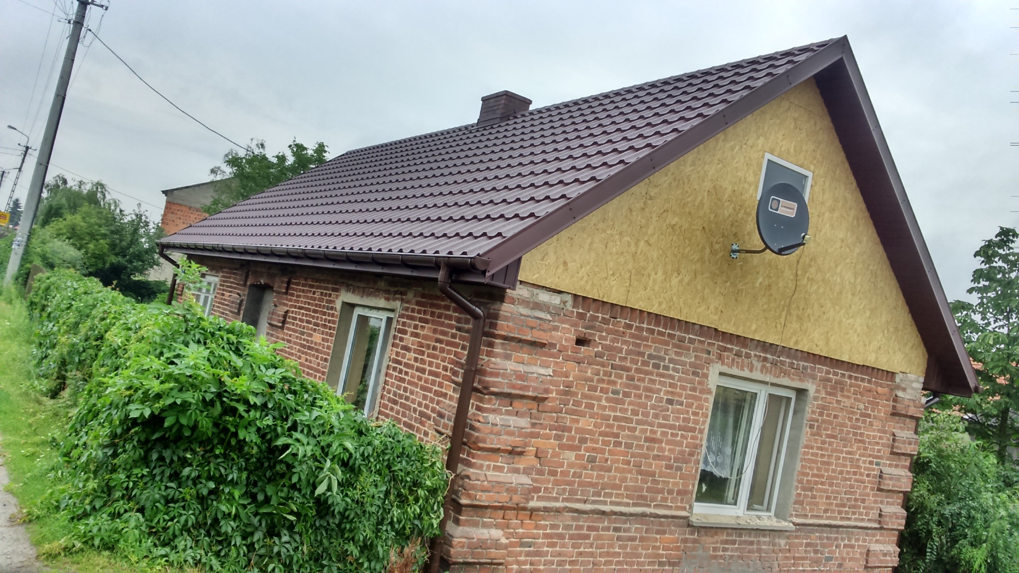 Dom z brązowym dachem z blachy trapezowej, obróbkami blacharskimi i ścianą szczytową z płyt OSB z zamontowaną anteną satelitarną.