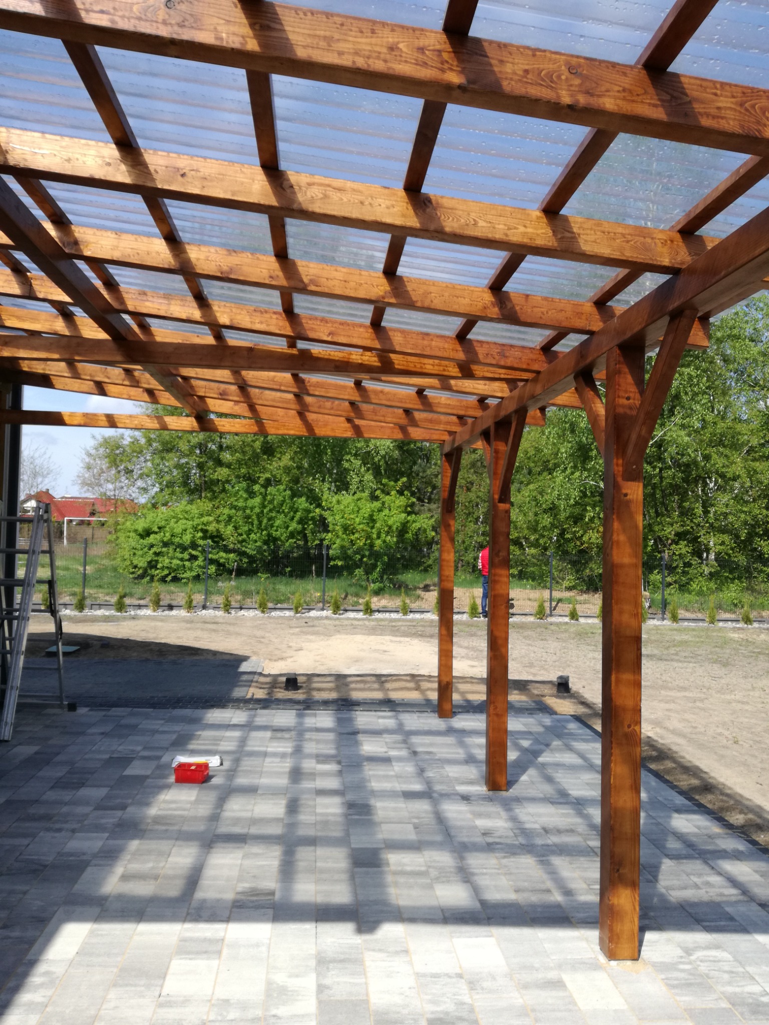 Drewniana konstrukcja z przezroczystym zadaszeniem, widoczne słupy, fragment kostki brukowej, drabina i osoba w oddali na tle zieleni.