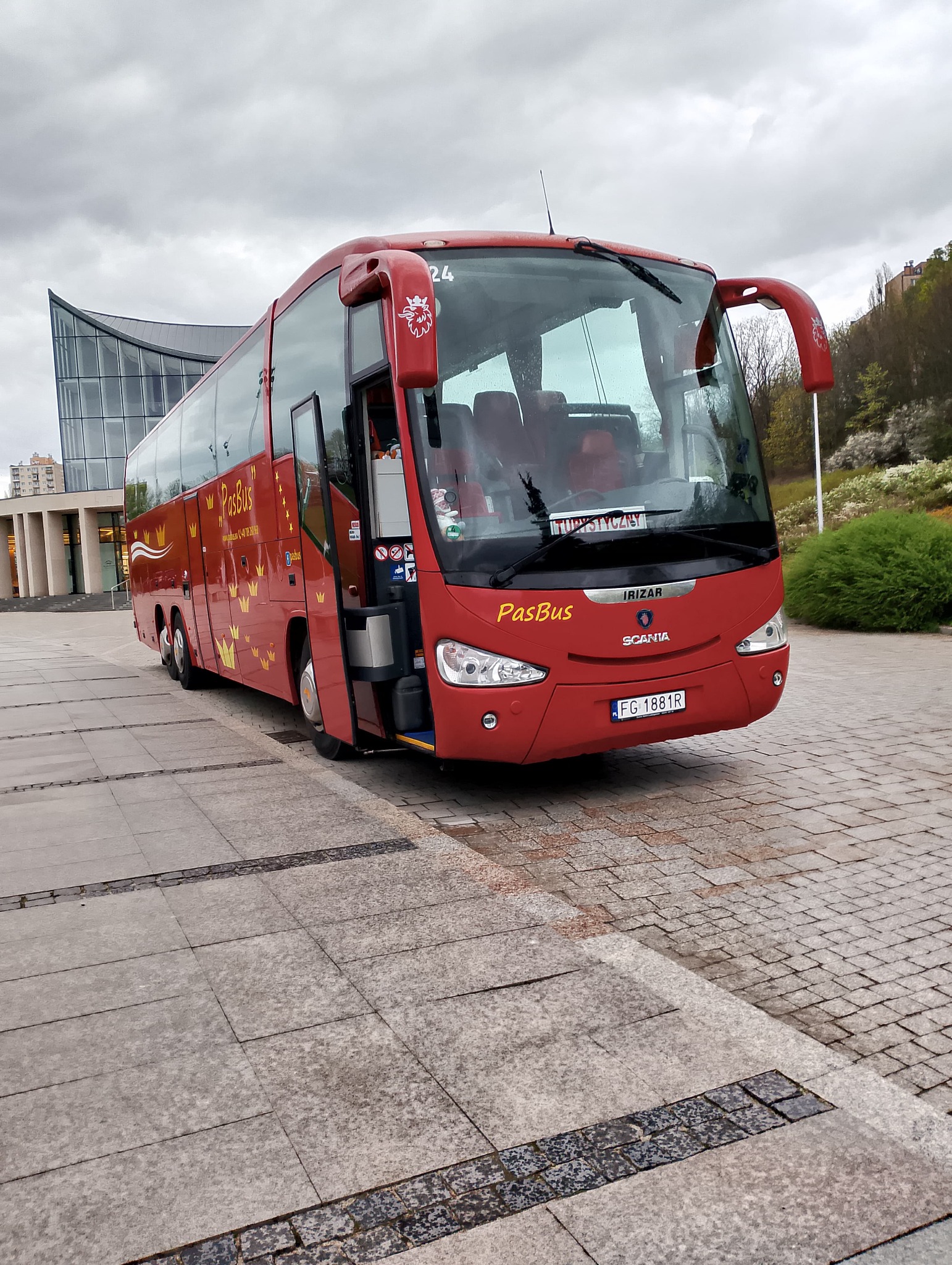 Czerwony autobus turystyczny PasBus marki Irizar Scania z otwartymi drzwiami stoi na placu przed nowoczesnym budynkiem o szklanej fasadzie.