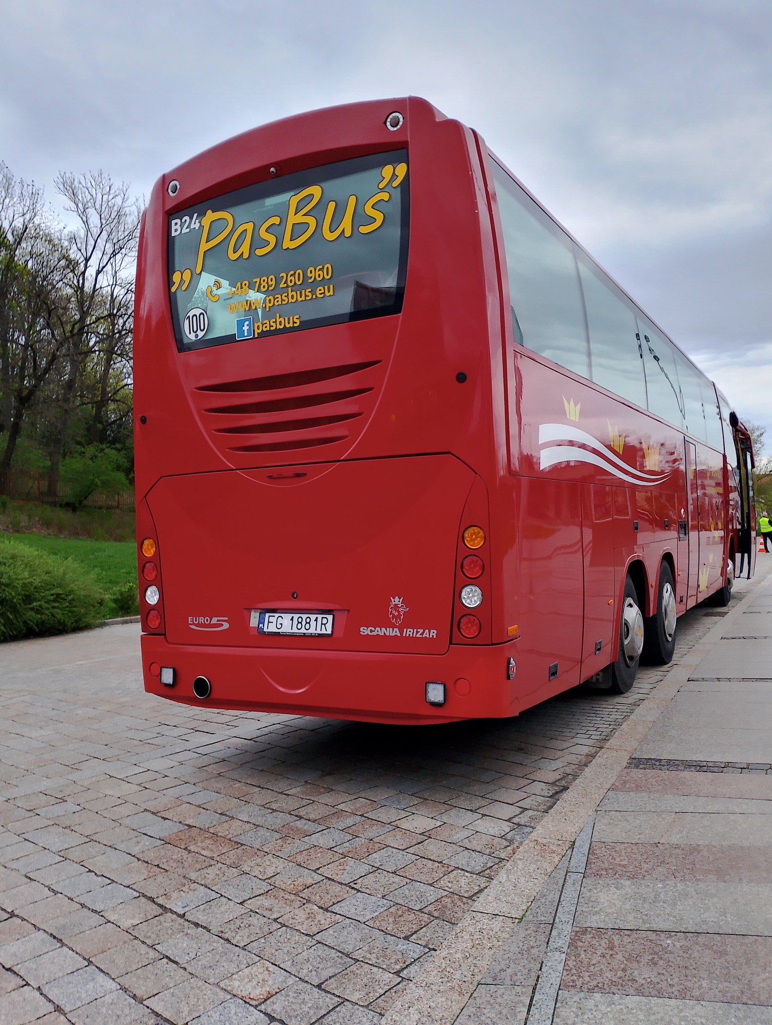 Tył czerwonego autobusu turystycznego marki Scania Irizar z żółtym logo firmy PasBus na szybie, zaparkowanego na brukowanym placu.