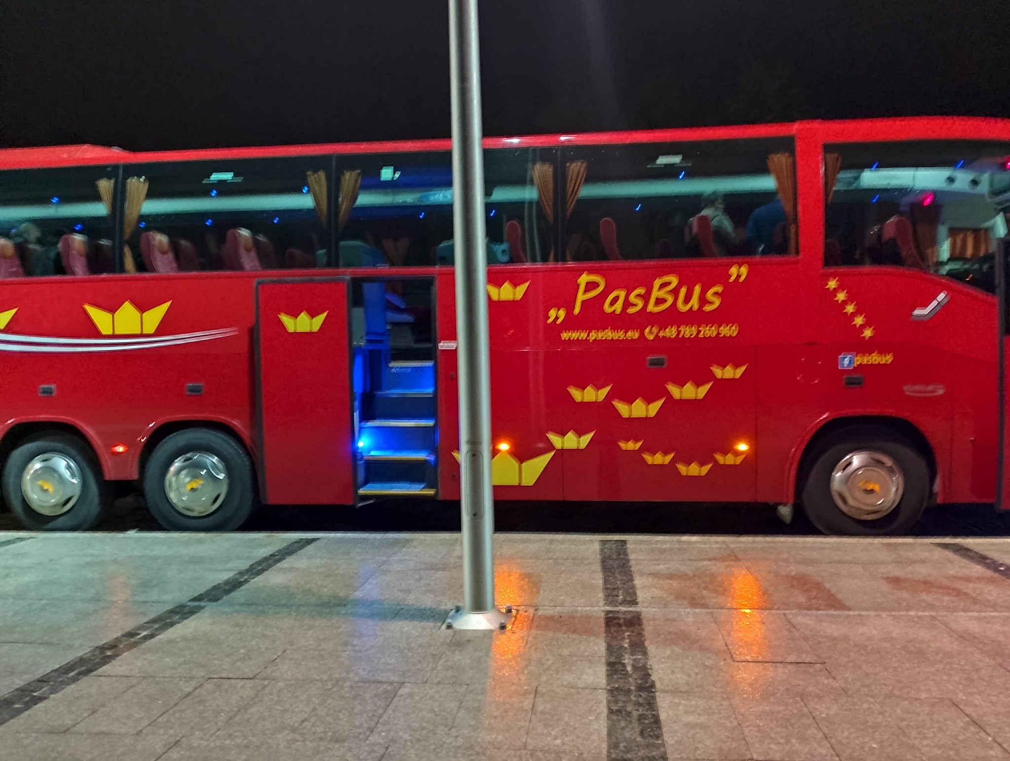 Czerwony autobus 'PasBus' z otwartymi drzwiami i podświetlonymi schodami na przystanku autobusowym w nocy. Autobus ozdobiony jest żółtymi koronami i posiada logo firmy.