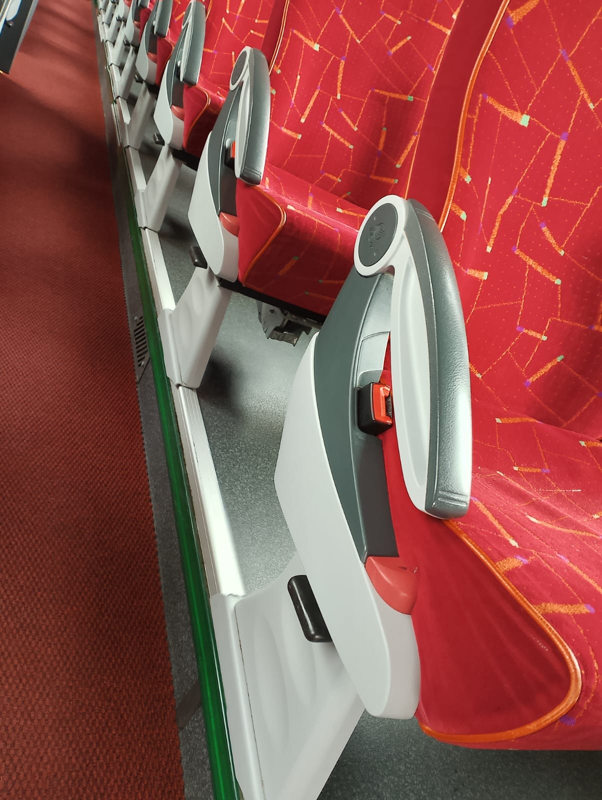 Rząd czerwonych, wzorzystych foteli z szarymi zagłówkami i pasami bezpieczeństwa w autobusie, widok z góry na bok.