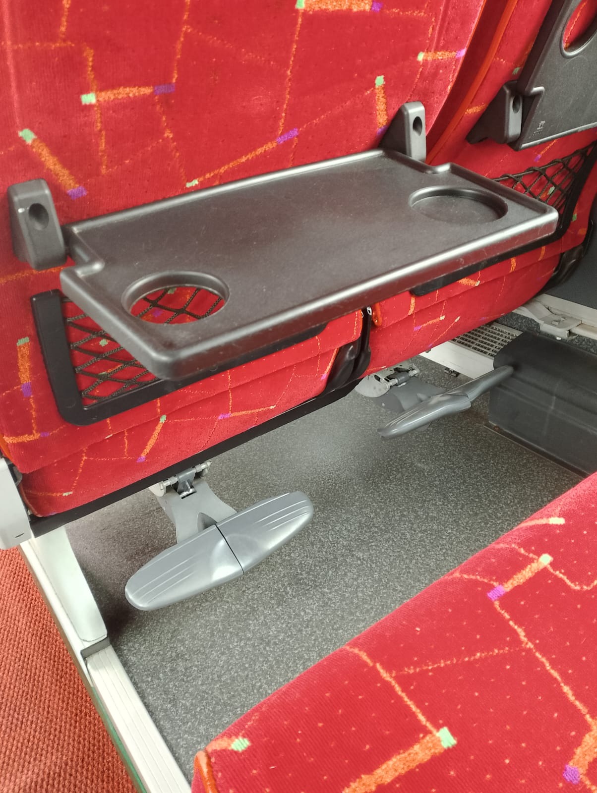 Wnętrze autobusu z czerwonymi siedzeniami z wzorami, rozkładanym stolikiem z uchwytem na napoje i podnóżkiem.