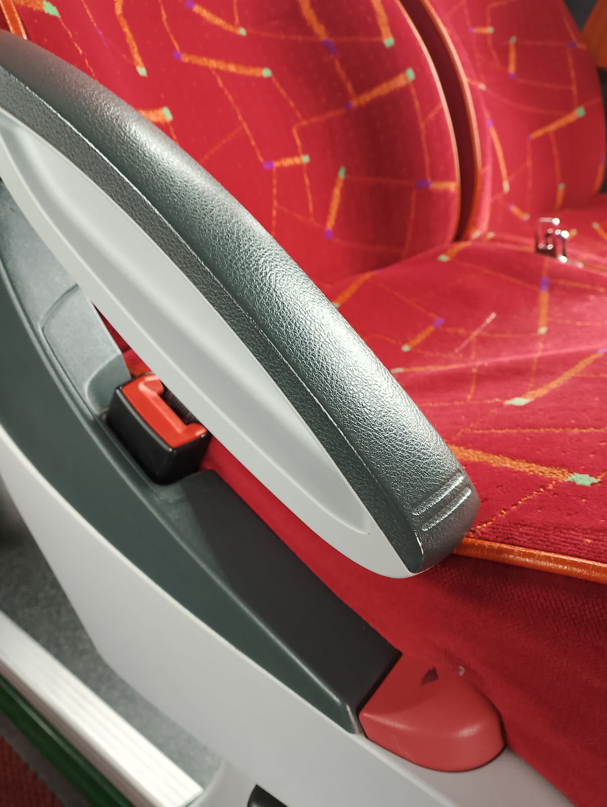 Szczegółowe ujęcie fotela pasażerskiego z podłokietnikiem i zapiętym pasem bezpieczeństwa, wzorzysta czerwona tapicerka.