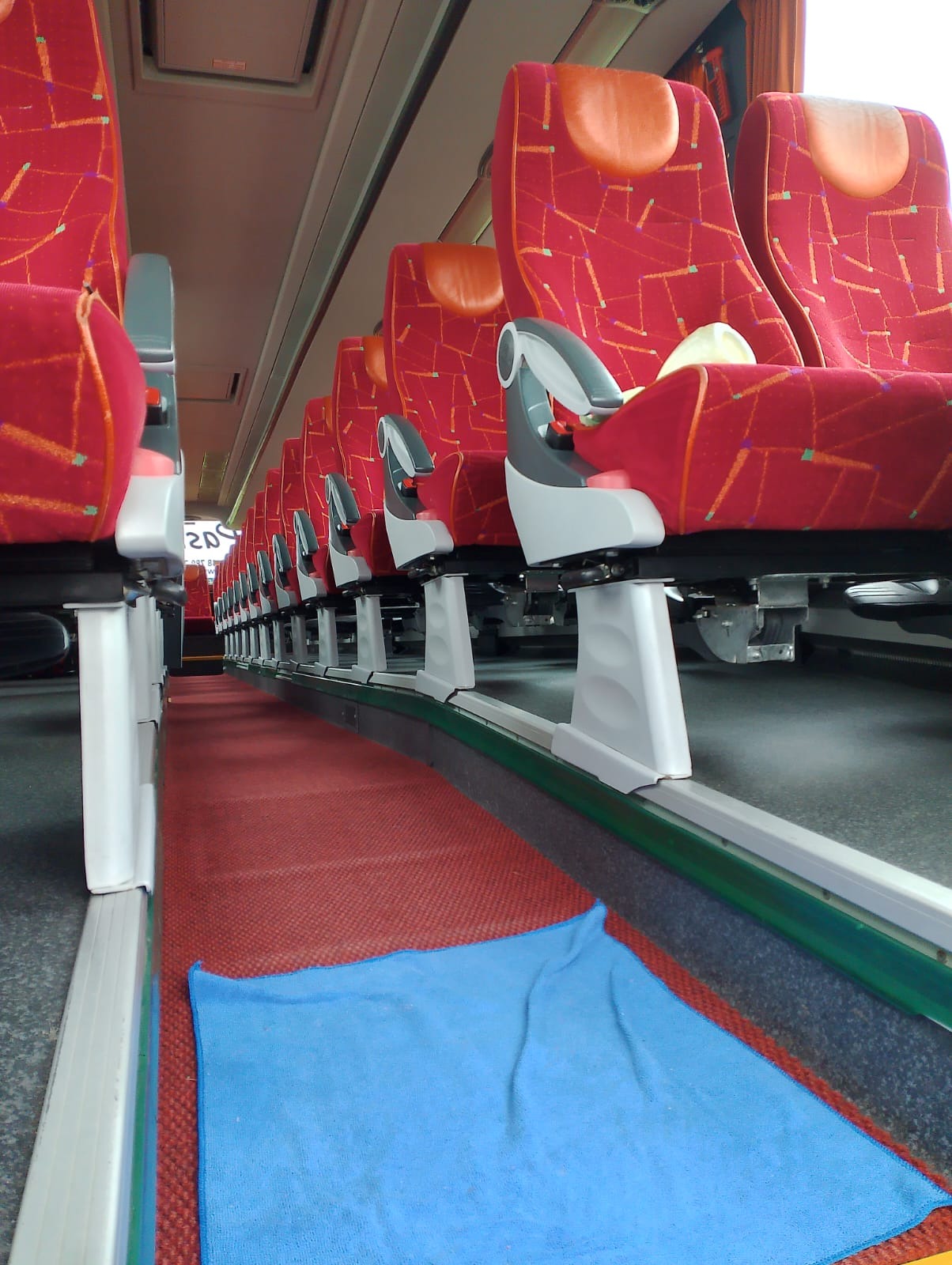 Wnętrze autobusu z rzędem czerwonych foteli z geometrycznym wzorem, szarymi podłokietnikami i niebieskim ręcznikiem na czerwonej wykładzinie w przejściu.