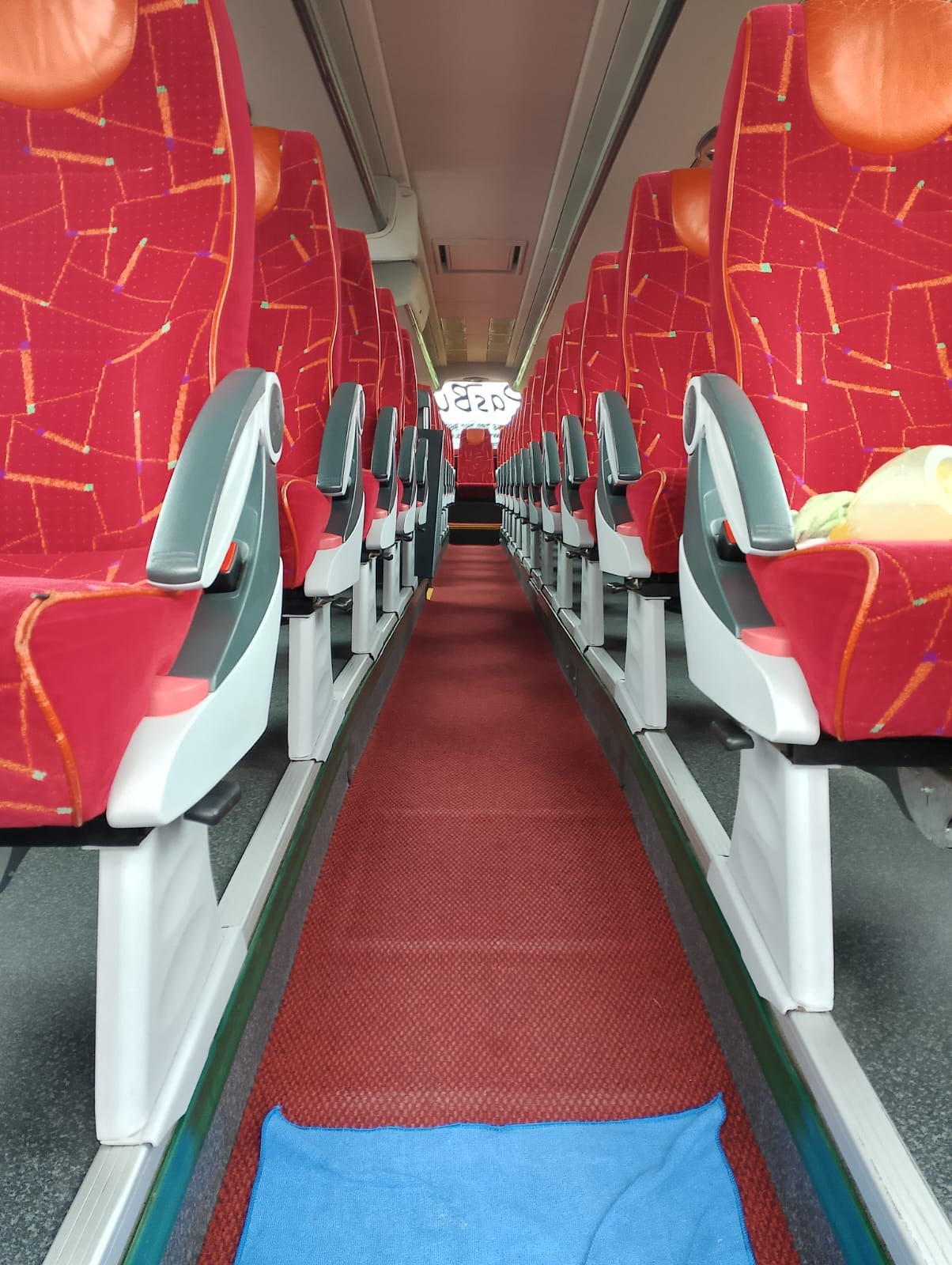 Wnętrze autobusu dalekobieżnego z czerwonymi fotelami z geometrycznym wzorem i czerwonym dywanem na podłodze, widok wzdłuż korytarza między rzędami siedzeń.