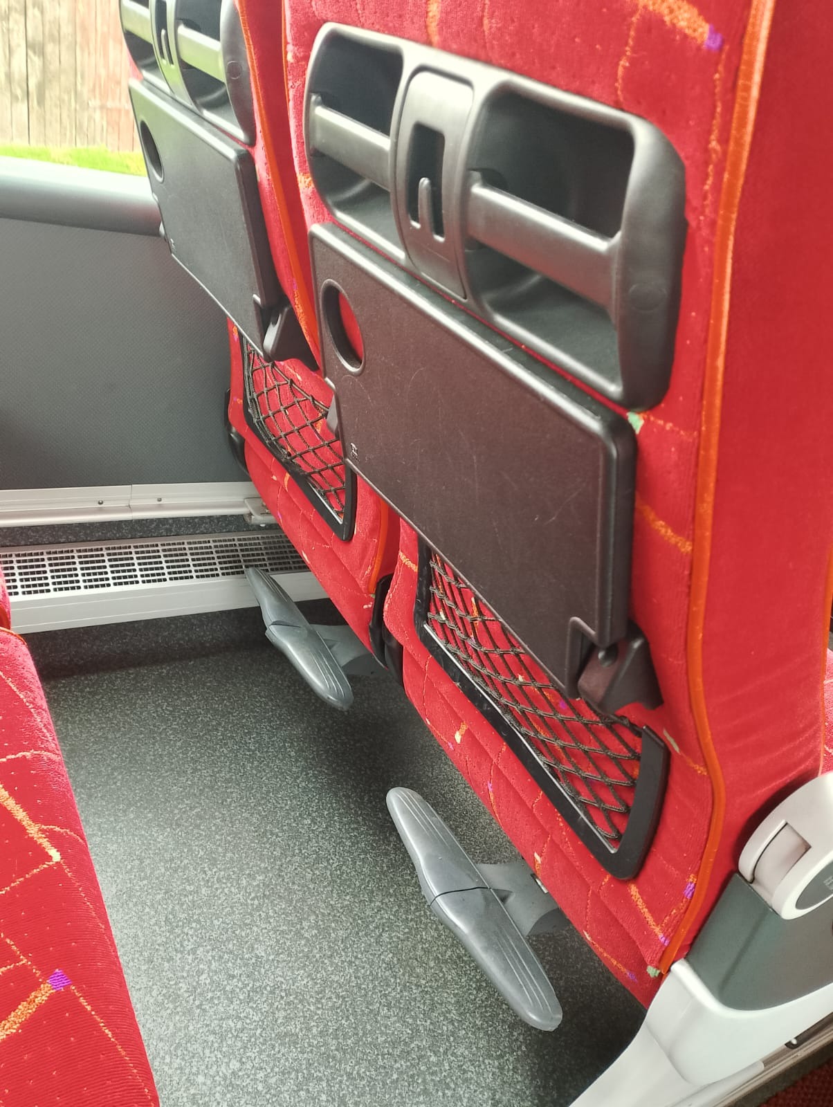 Wnętrze autobusu: czerwone fotele z rozkładanymi stolikami i siatkami na drobiazgi, widoczne podnóżki i kratka wentylacyjna.