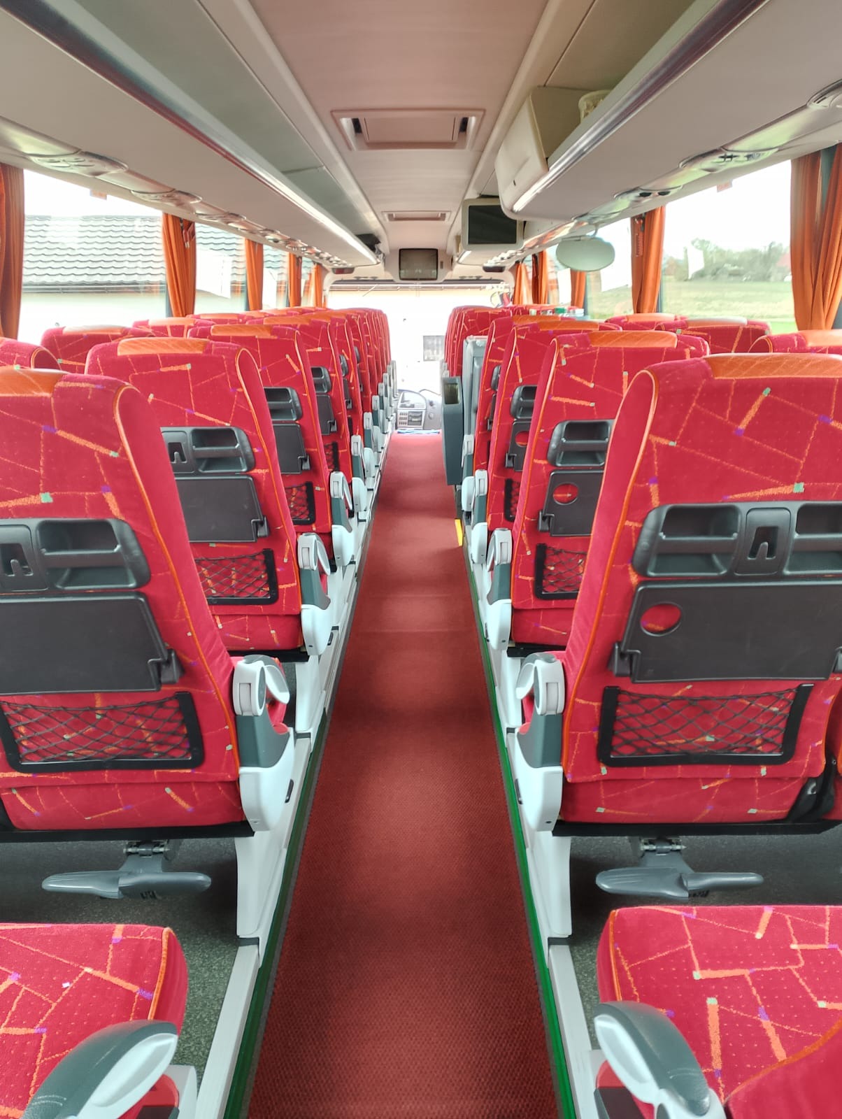 Wnętrze autobusu turystycznego z czerwonymi, wzorzystymi siedzeniami, rozkładanymi stolikami i siatkami na bagaż, widok na przejście i przednią szybę z perspektywy pasażera.