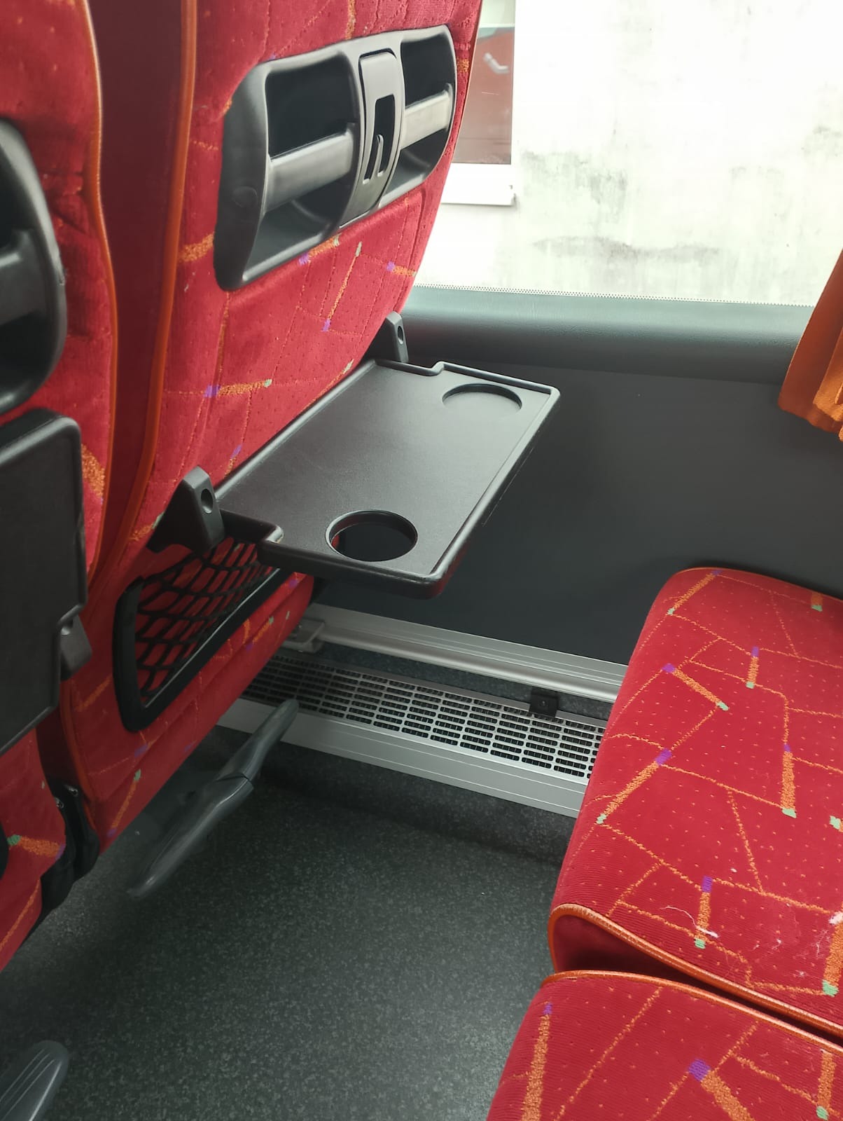 Wnętrze autobusu: czerwone fotele z rozkładanym, czarnym stolikiem z wgłębieniami na napoje, widoczna kratka wentylacyjna pod oknem.