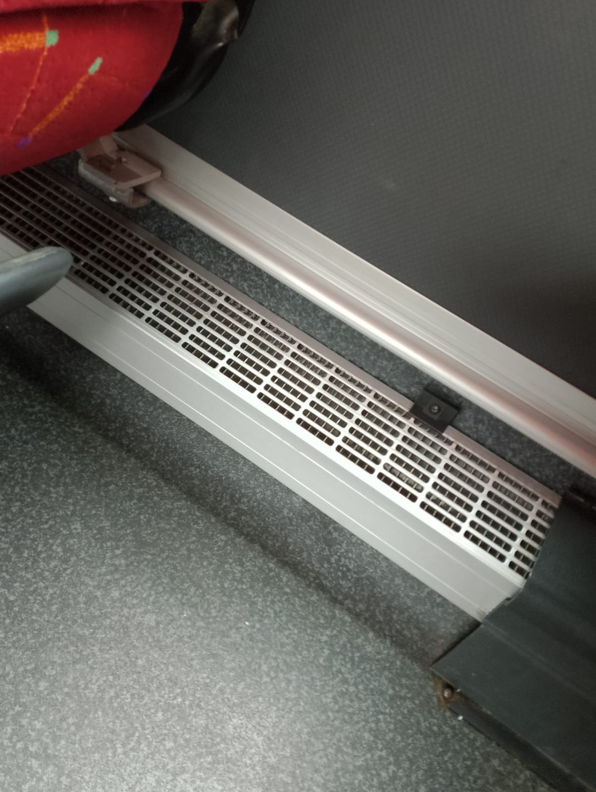 Wnętrze autobusu: szara podłoga, fragment czerwonego siedzenia i srebrna kratka wentylacyjna wzdłuż ściany.