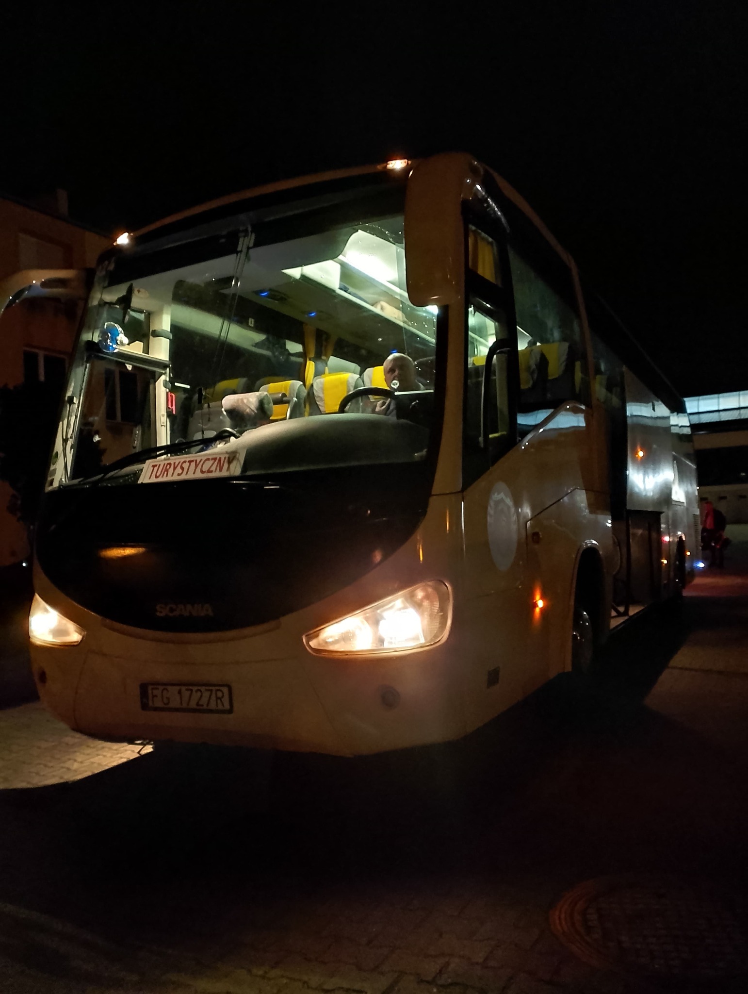 Żółty autokar turystyczny Scania z włączonymi światłami, zaparkowany nocą, widoczny kierowca wewnątrz.