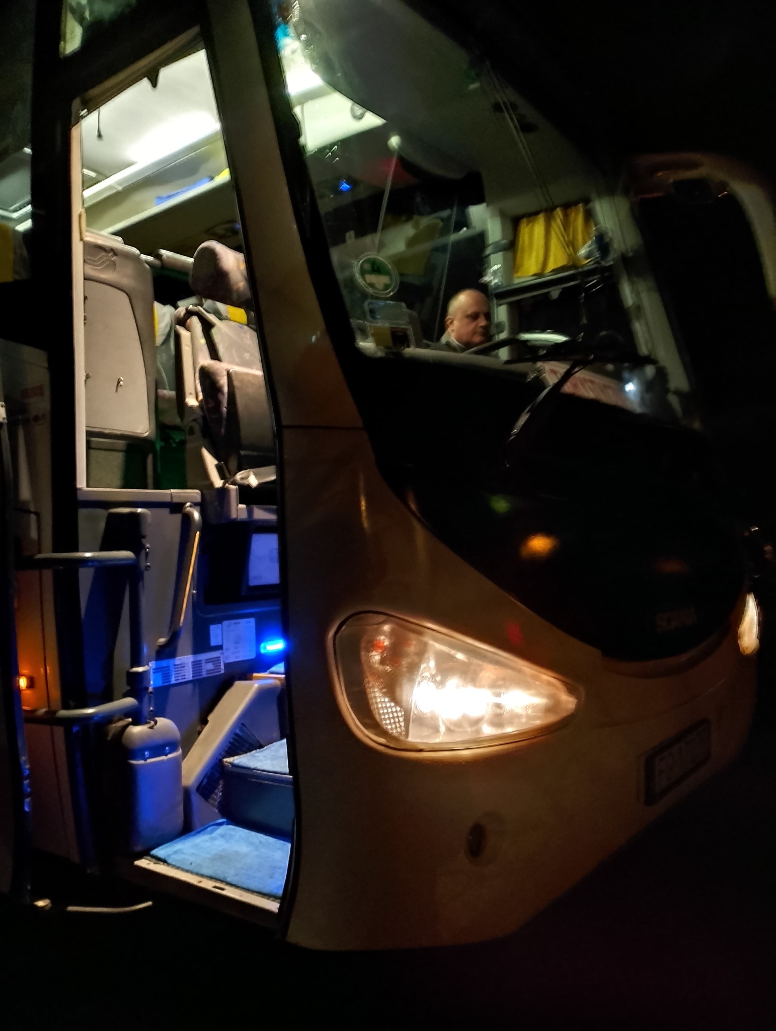 Oświetlony przód autobusu z widocznymi schodkami wejściowymi wyłożonymi niebieską wykładziną i kierowcą za kierownicą w nocy.