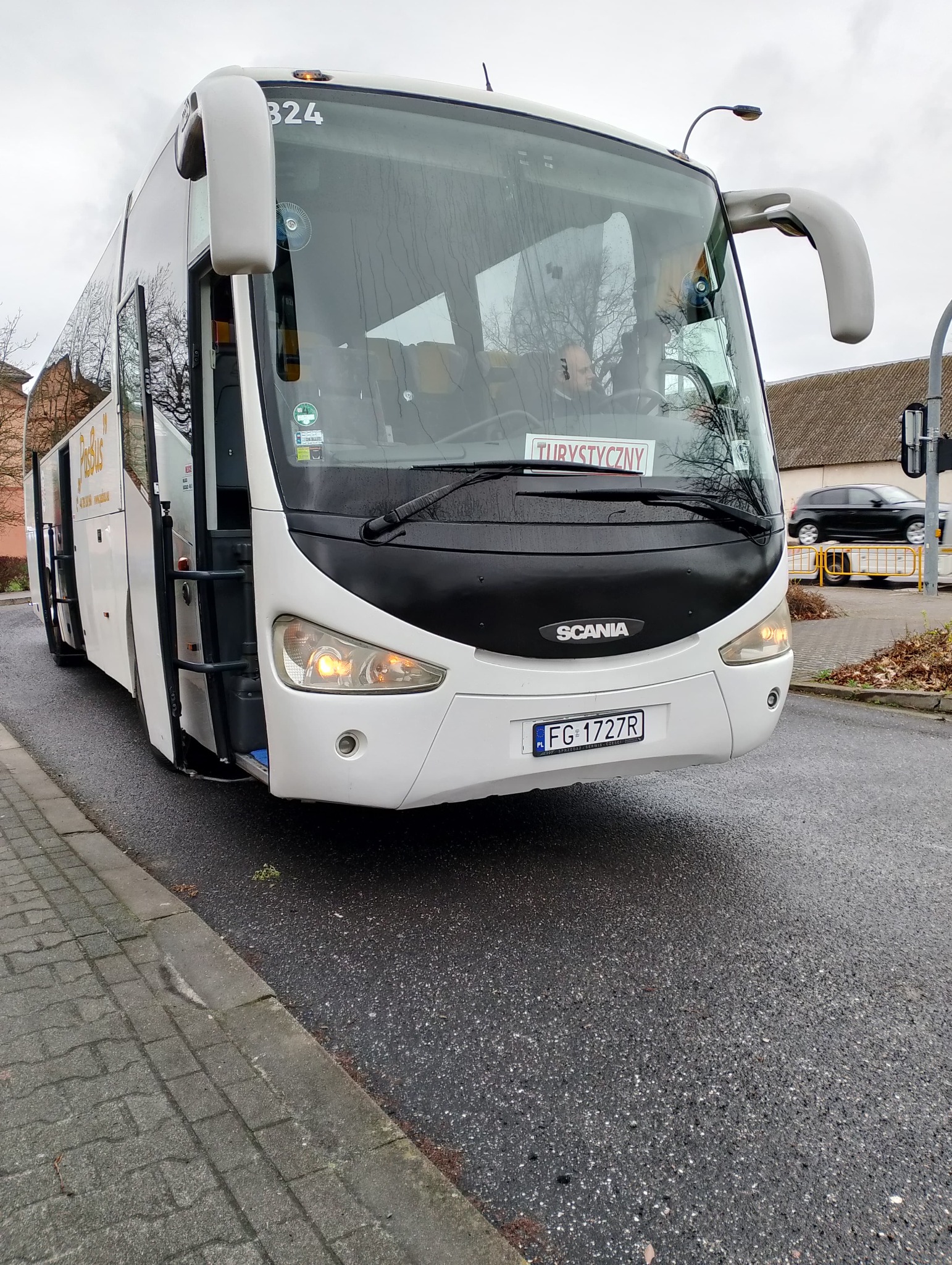 Biały autokar Scania z otwartymi drzwiami na mokrej jezdni, tablica 'Turystyczny' za szybą, widoczny kierowca.