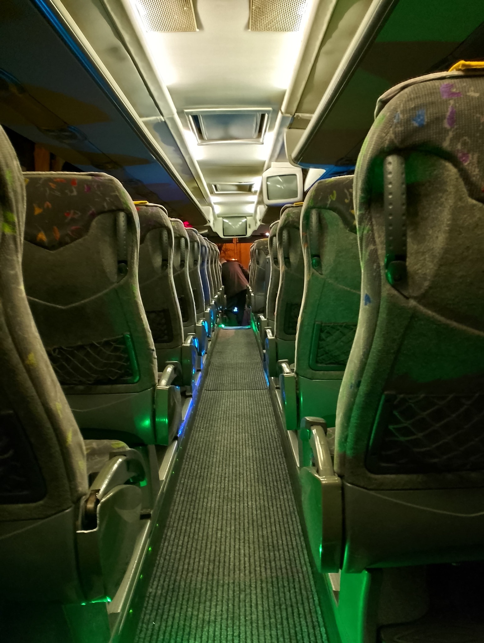 Wnętrze autobusu z rzędem foteli z szarymi tapicerkami w geometryczne wzory, oświetlonych zielonymi diodami LED wzdłuż korytarza, z widocznym pasażerem idącym w stronę tyłu pojazdu.