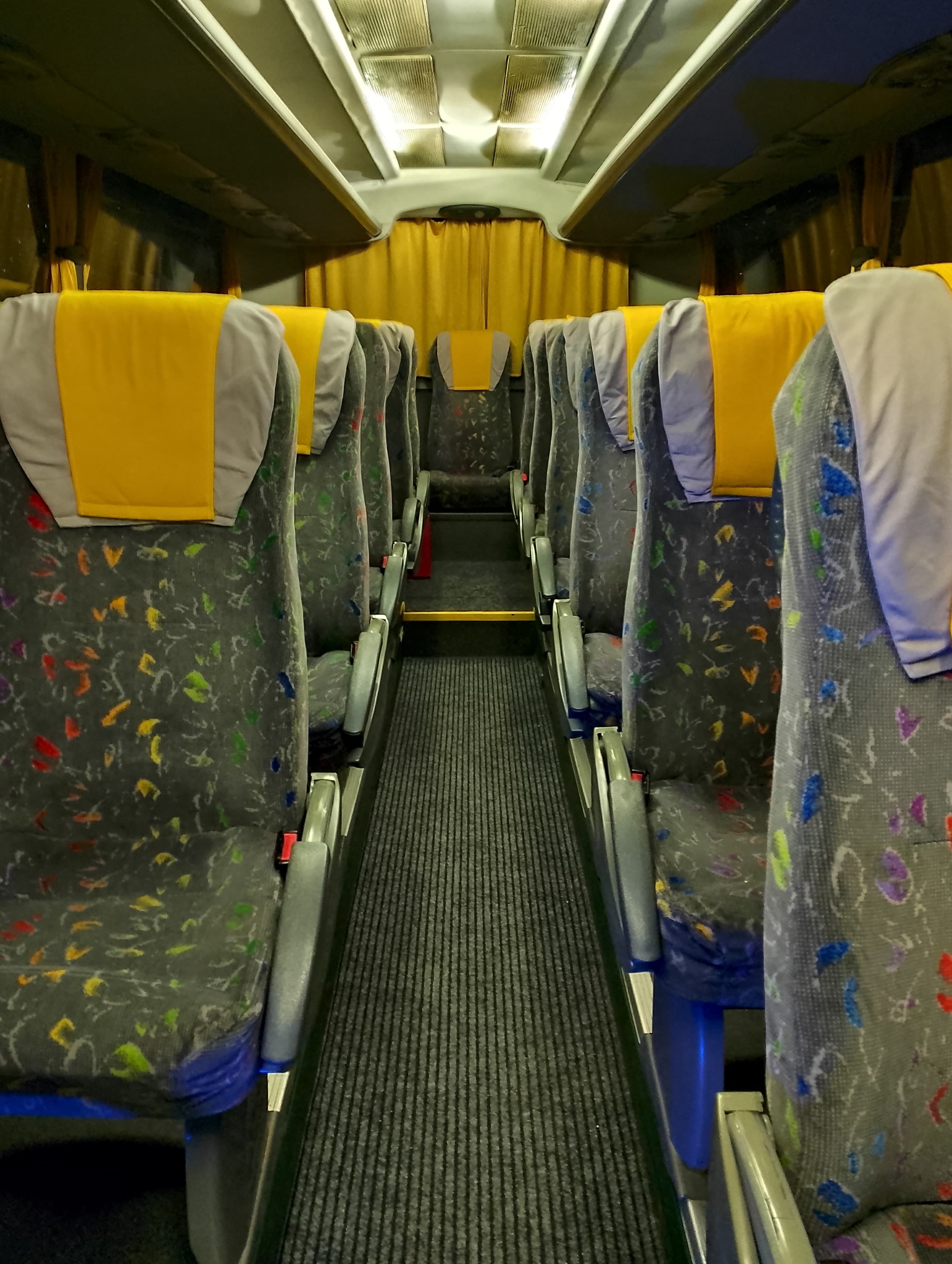 Wnętrze autobusu z rzędami foteli z szarą tapicerką w kolorowe wzory i żółtymi zagłówkami, widok wzdłuż korytarza z szarą wykładziną.