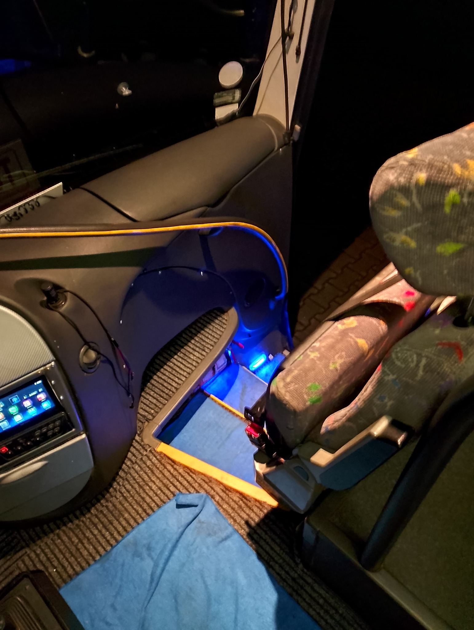 Wnętrze autobusu z podświetleniem LED, widoczne fotele z kolorowym wzorem, panel sterowania i fragment niebieskiej szmatki na podłodze.