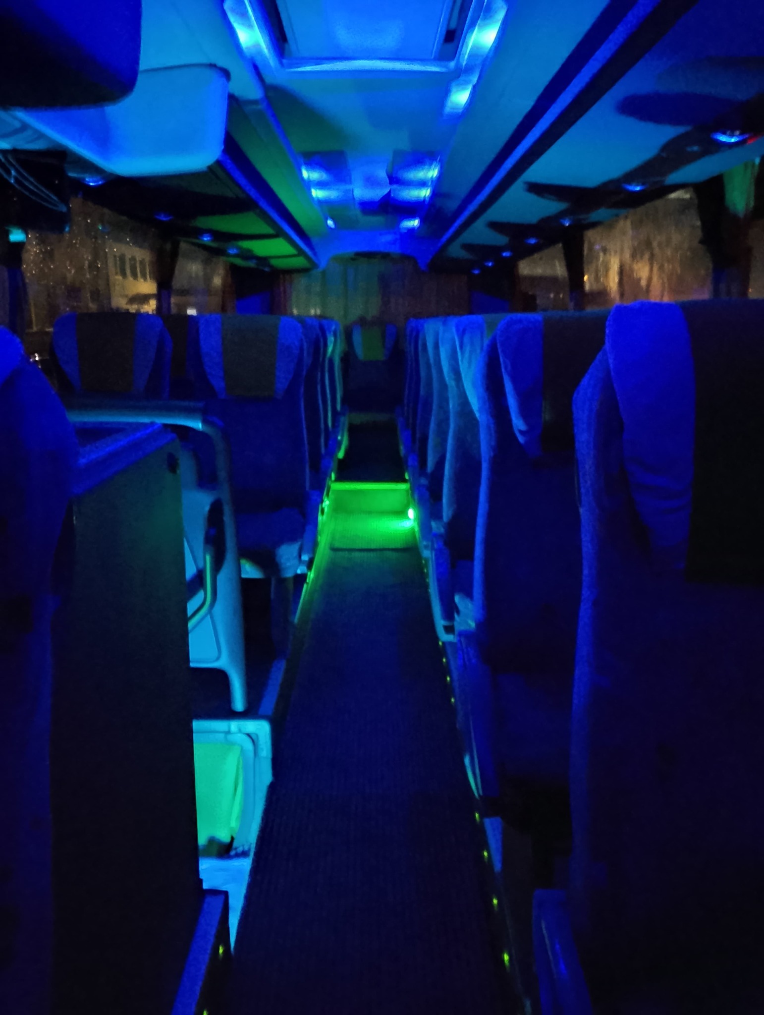 Wnętrze autobusu oświetlone niebieskim i zielonym światłem LED, puste fotele z ciemnymi zagłówkami, widok na korytarz wzdłuż rzędów siedzeń.