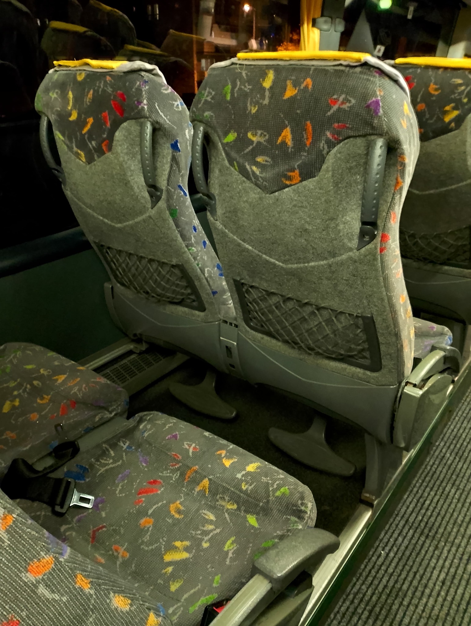 Wnętrze autobusu z szarymi fotelami w kolorowe wzory, widoczny pas bezpieczeństwa i siatka na oparciu fotela.