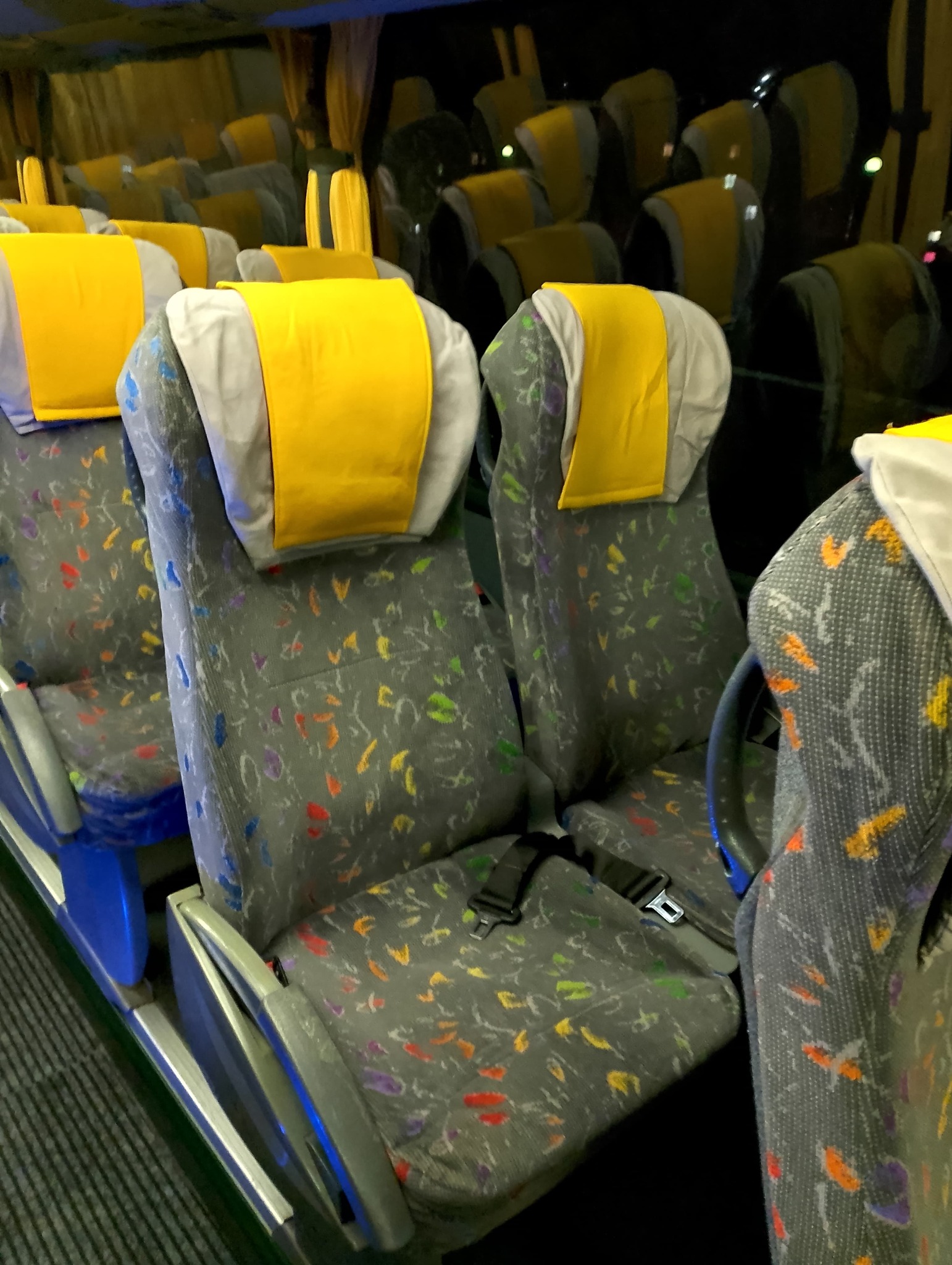 Wnętrze autobusu z rzędami foteli obitych szarą tkaniną w kolorowe wzory, z żółtymi zagłówkami i czarnymi pasami bezpieczeństwa.