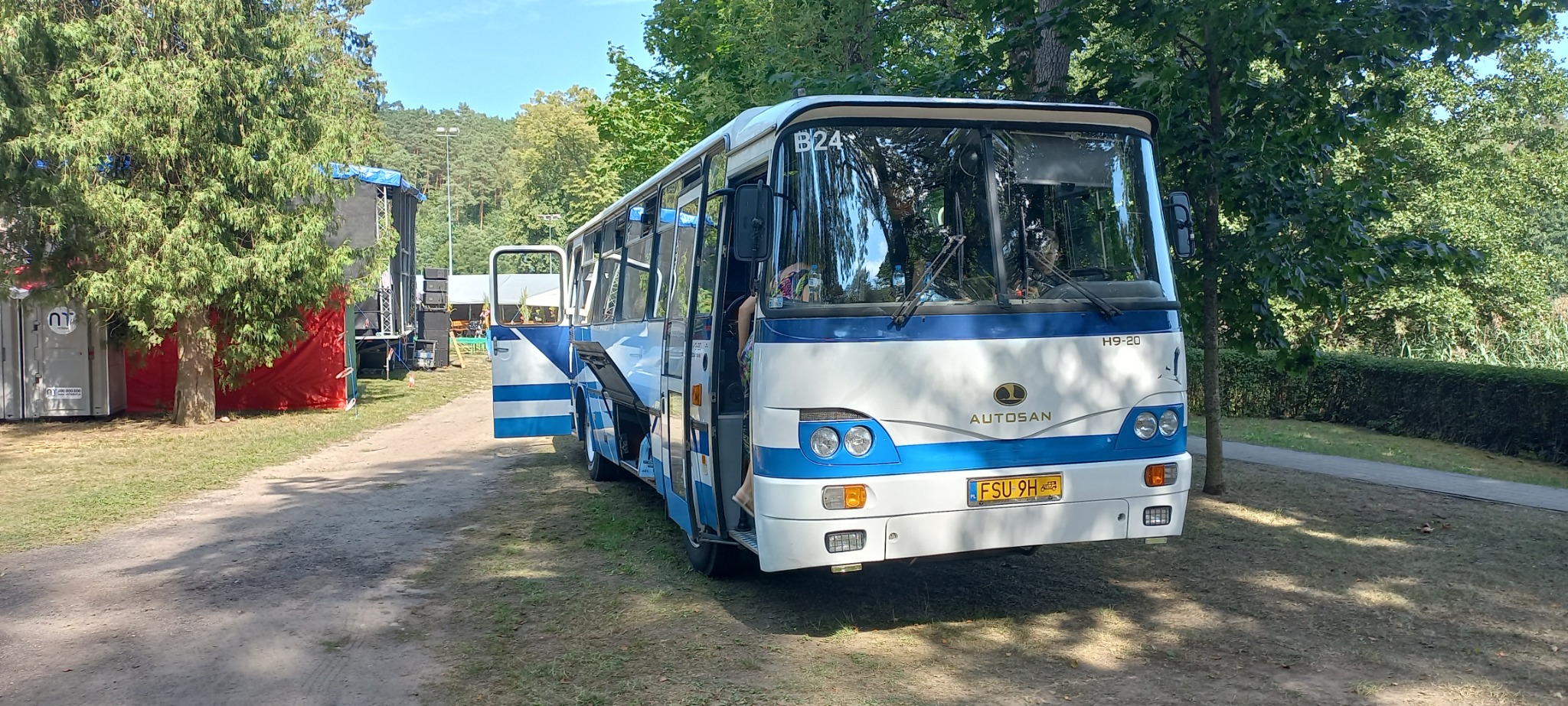 Autosan H9-20 w barwach biało-niebieskich z otwartymi drzwiami, zaparkowany na terenie zielonym z widoczną sceną w tle.