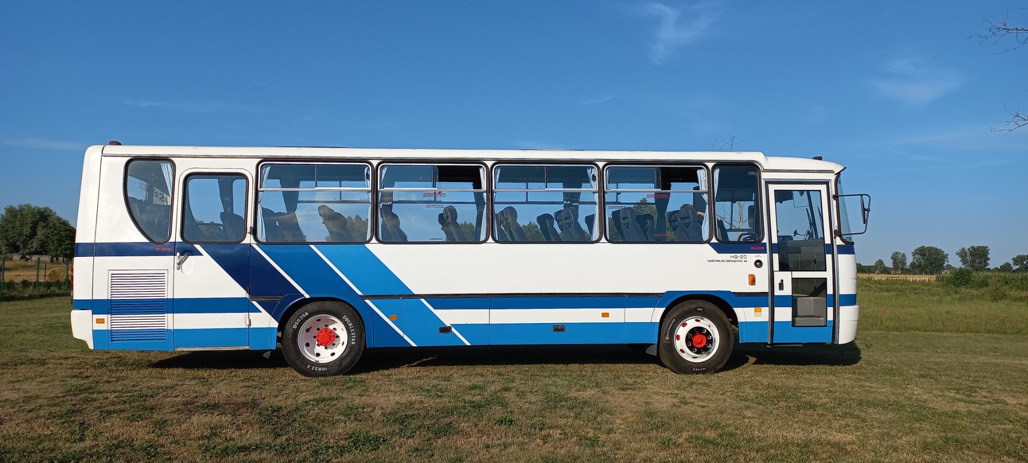 Biało-niebieski autobus miejski Autosan H9-20 z otwartymi drzwiami, stojący na trawiastym polu w słoneczny dzień.