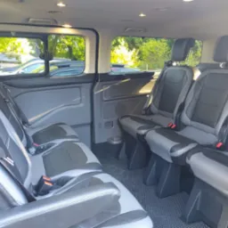 Wnętrze przestronnego busa pasażerskiego z trzema rzędami foteli w odcieniach szarości i czerni, widoczne przez otwarte drzwi przesuwne.