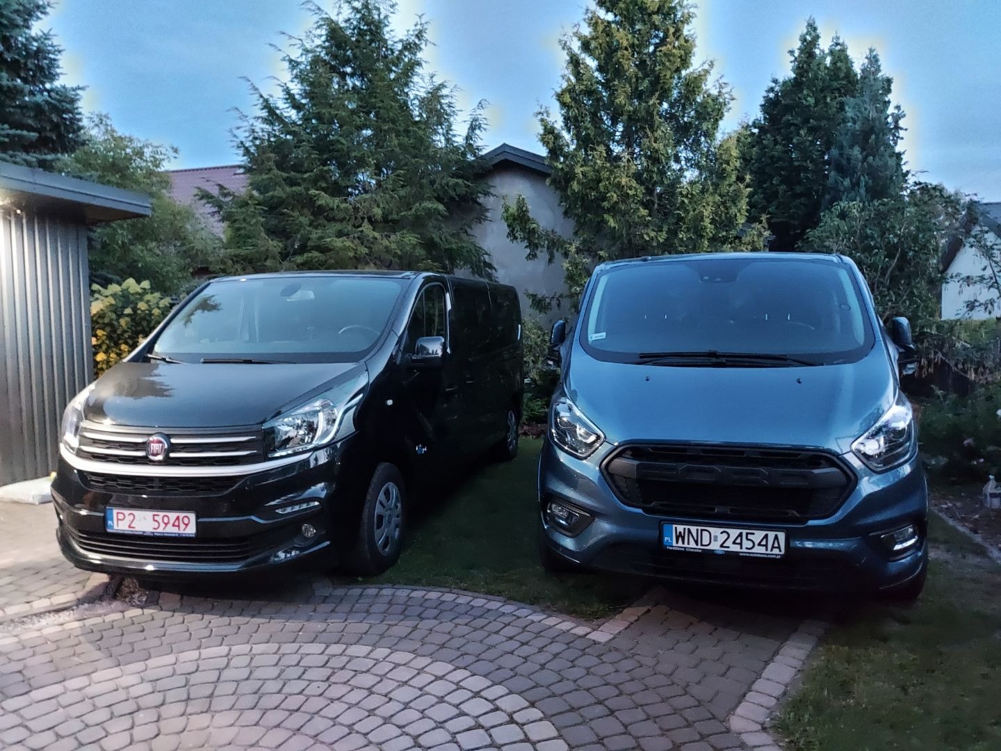 Dwa busy: czarny Fiat i niebieski Ford Transit, zaparkowane na posesji z kostki brukowej, w tle drzewa i budynek.