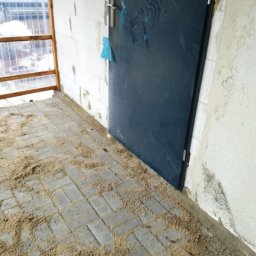 HydroBrux - Szare kostki brukowe ułożone na tarasie w Kraśniku, zasypane piaskiem, widok na ścianę budynku i drzwi wejściowe.