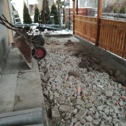 HydroBrux - Przygotowanie podłoża pod kostkę: gruz i kamienie na gruncie obok drewnianego płotu, taczka oparta o ścianę budynku.