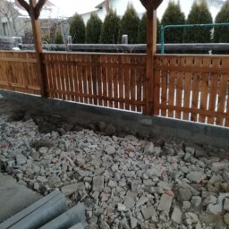 HydroBrux - Drewniane ogrodzenie panelowe z widocznymi słupkami, częściowo ukończone, z gruzem i elementami betonowymi w tle, sugerujące prace przygotowawcze.