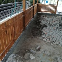 HydroBrux - Przygotowanie podłoża pod taras z drewnianą balustradą, widoczne warstwy ziemi, gruzu i fundament z bloczków betonowych.