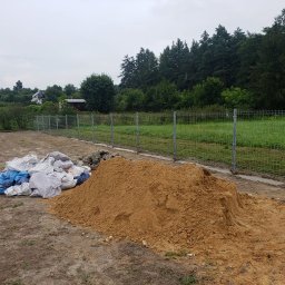 HydroBrux - Stos piasku budowlanego i worki obok nowo postawionego ogrodzenia z siatki na tle zieleni.