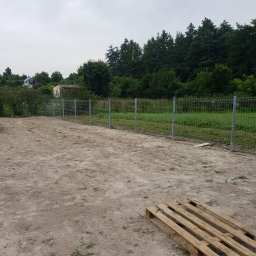 HydroBrux - Nowo postawione ogrodzenie z siatki na metalowych słupkach, widoczne na piaszczystym terenie z paletą w rogu, w tle zieleń drzew i krzewów pod pochmurnym niebem.