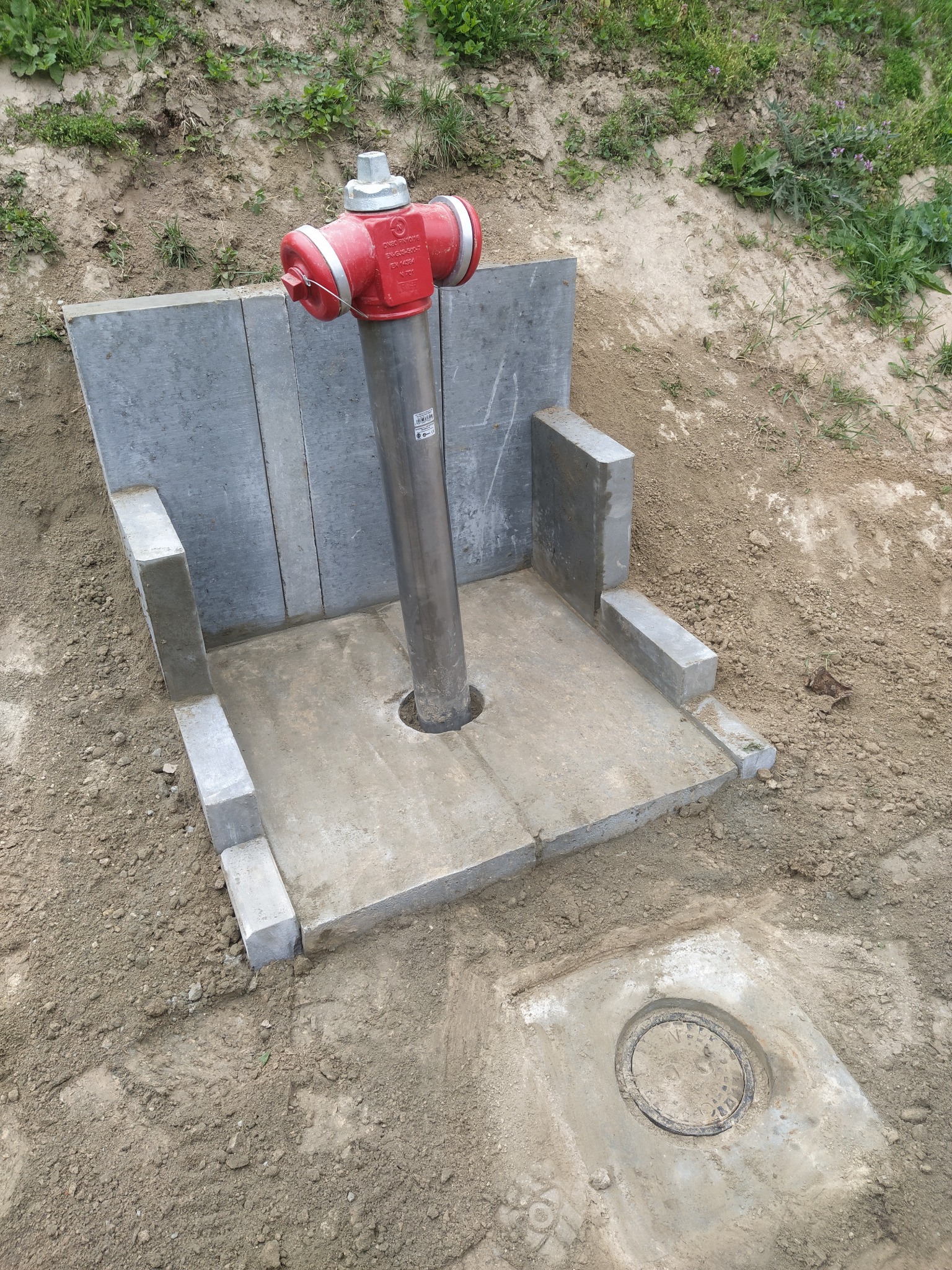 Czerwony hydrant przeciwpożarowy osadzony w betonowej obudowie z szarymi płytami, widoczne przyłącze kanalizacyjne w ziemi.