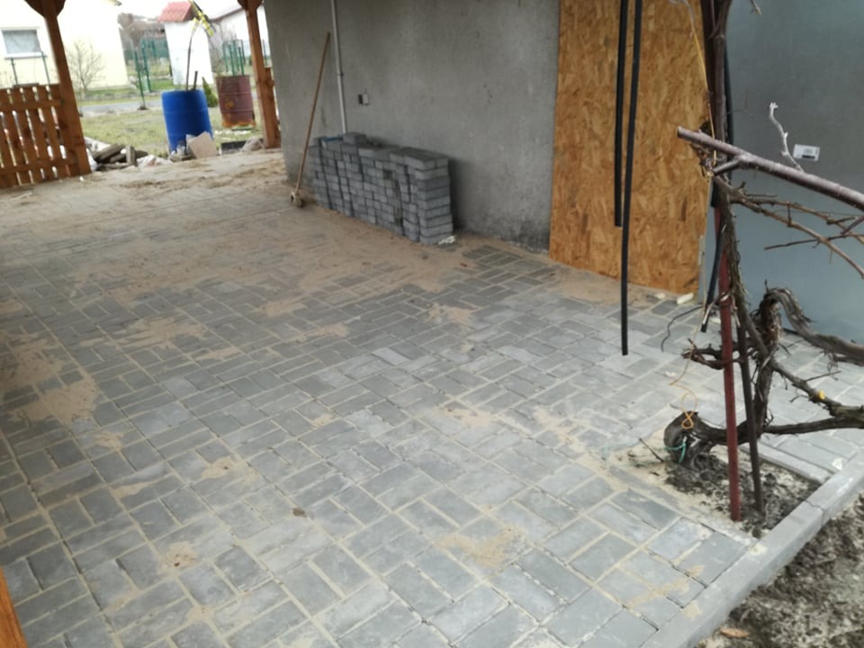 Ułożona szara kostka brukowa na podwórku, z widocznym stosem kostki do dalszej pracy i rozłożonym piaskiem na powierzchni, częściowo zadaszone miejsce.