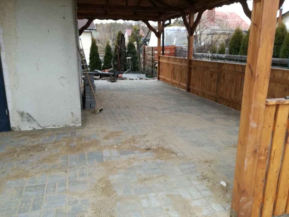 Szara kostka brukowa ułożona pod drewnianą wiatą, zasypana piaskiem po fugowaniu, widoczne narzędzia i materiały budowlane, w tle ogrodzenie i roślinność.