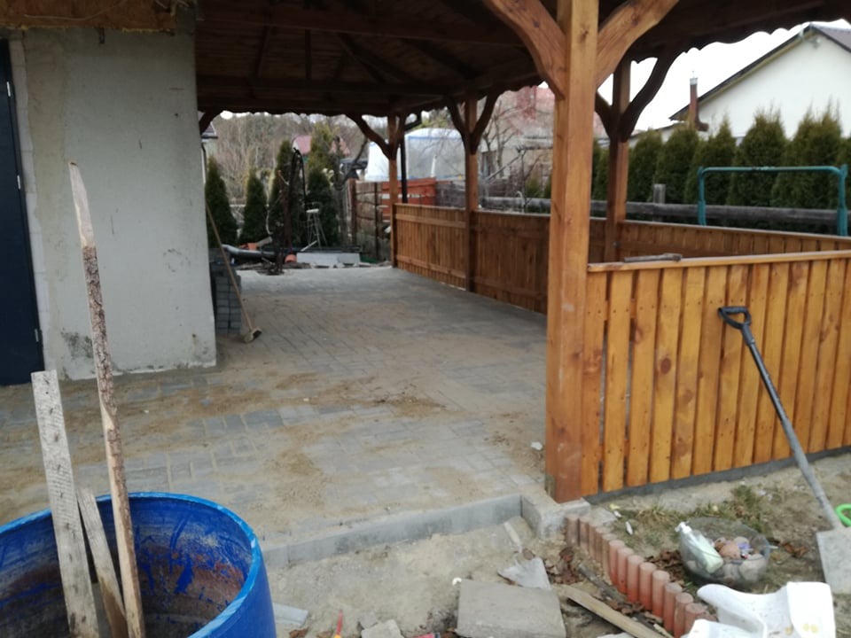 Ułożona szara kostka brukowa pod drewnianą wiatą z drewnianą balustradą, widoczne narzędzia i materiały budowlane, etap prac wykończeniowych.