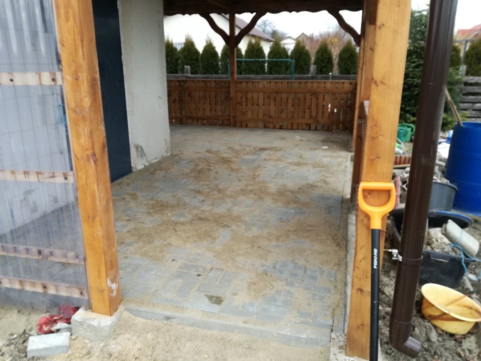 Zadaszona przestrzeń z ułożoną szarą kostką brukową, widoczna łopata Fiskars oparta o drewniany słup, w tle drewniana balustrada i równe tuje.