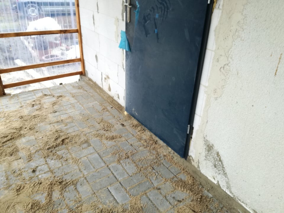 Szare kostki brukowe ułożone na tarasie w Kraśniku, zasypane piaskiem, widok na ścianę budynku i drzwi wejściowe.