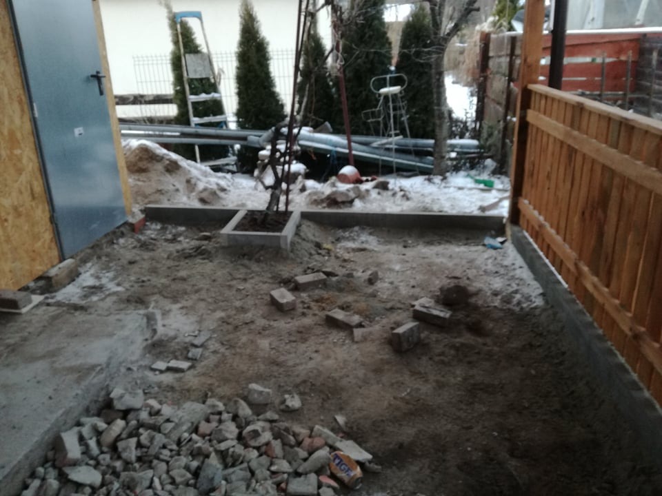 Przygotowanie podłoża pod kostkę brukową w ogrodzie, widoczne obrzeża, gruz ceglany, ziemia i częściowo drewniany płot oraz śnieg.