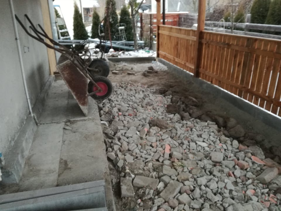 Przygotowanie podłoża pod kostkę: gruz i kamienie na gruncie obok drewnianego płotu, taczka oparta o ścianę budynku.