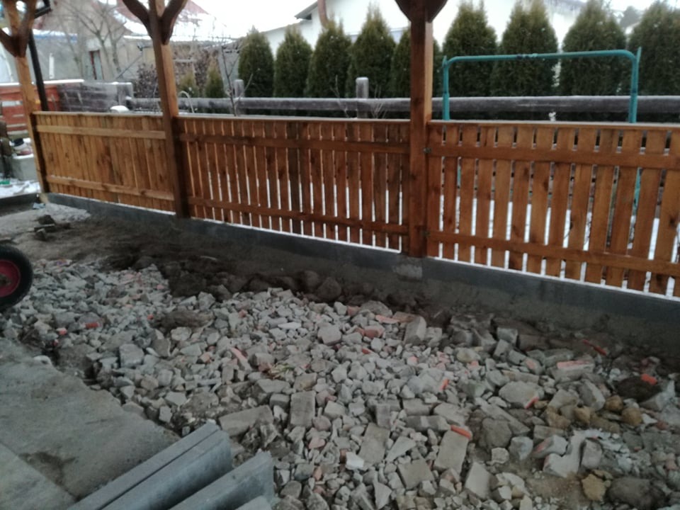 Drewniane ogrodzenie panelowe z widocznymi słupkami, częściowo ukończone, z gruzem i elementami betonowymi w tle, sugerujące prace przygotowawcze.