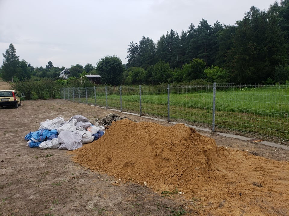 Stos piasku budowlanego i worki obok nowo postawionego ogrodzenia z siatki na tle zieleni.